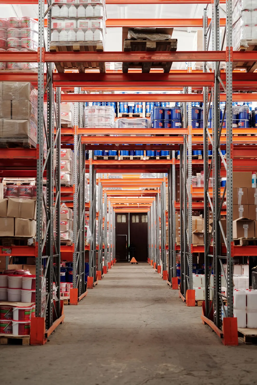 Long warehouse aisle between tall red pallet racks holding boxes and drums